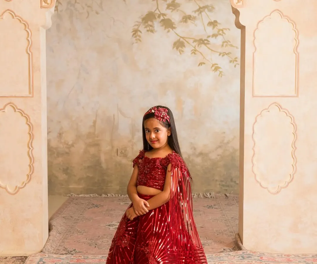 A classic red lehenga is a must have for any festive wardrobe. This custom stitched piece features shimmering sequin work and a delicate cape, ensuring she stands out at any wedding or celebration. We can customize this design in any color you choose.