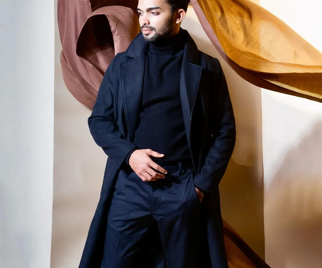 A full-length portrait of a male model in a sophisticated, dark navy trench coat and matching turtleneck. The studio shot uses draped brown and gold fabrics in the background to create an artistic, high-fashion mood for this menswear campaign.
