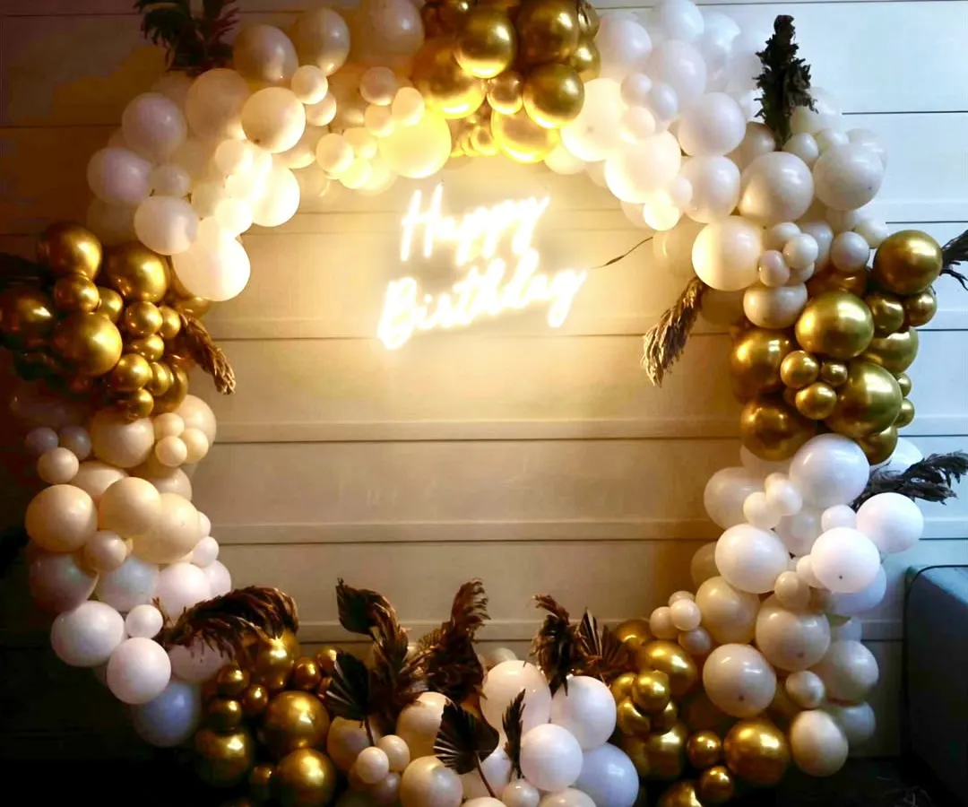 Elegance in every detail. This sophisticated birthday setup features a beautiful ring of white and gold chrome balloons, accented with delicate pampas grass and a warm 'Happy Birthday' neon sign for a touch of modern class.