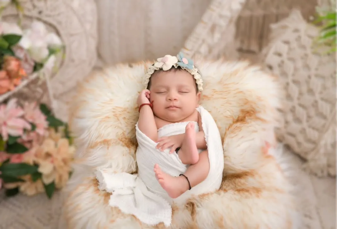 A peaceful newborn rests on a soft, cream-colored fur, adorned with a delicate floral headband. This photograph highlights our skill in posed newborn artistry, creating serene, beautiful portraits that focus on your baby's innocent features against a gentle, boho-inspired backdrop.