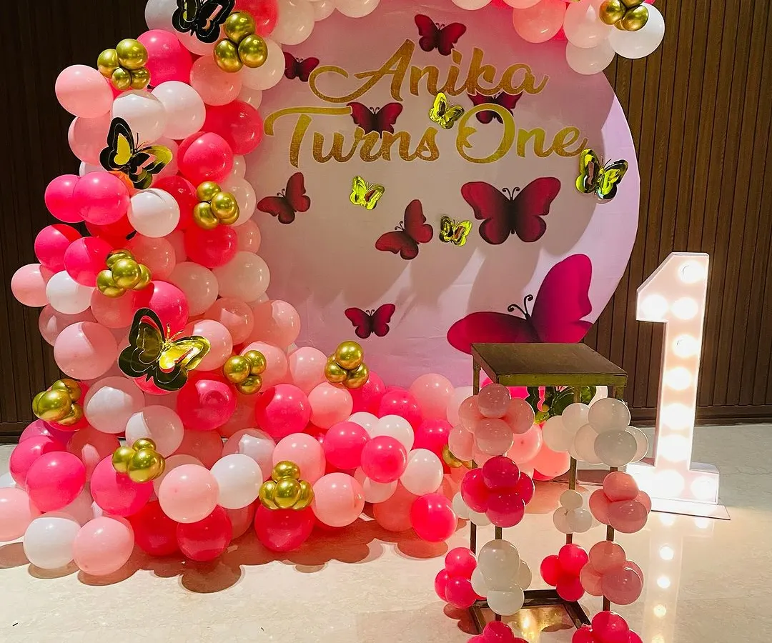 This is a popular butterfly theme decoration I did for a first birthday. It features a dense balloon arch in shades of pink and white, accented with gold and pink butterflies, and a large light-up marquee number '1'.