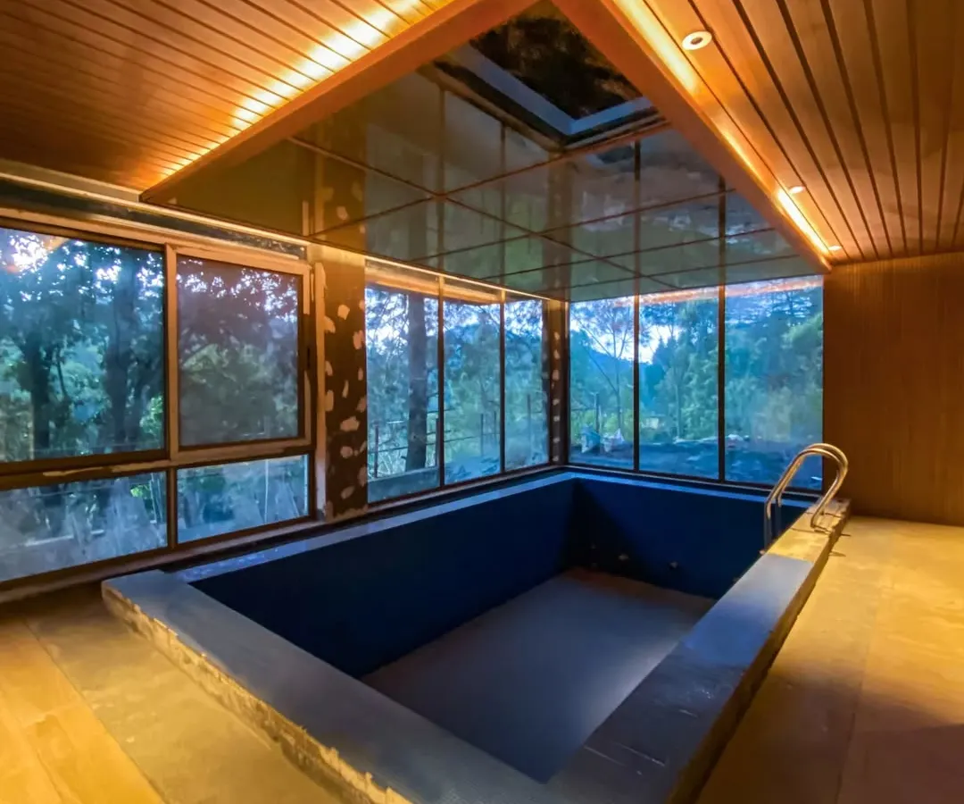 An indoor heated pool designed with a glass ceiling and panoramic windows, blurring the line between the interior and the lush Uttarakhand landscape. This space exemplifies our commitment to luxury design that incorporates natural light and views.