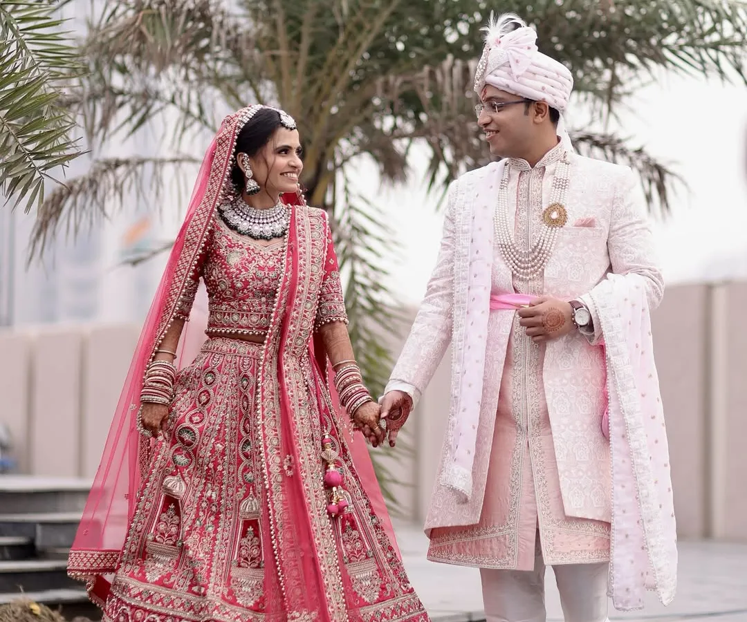 A candid shot of a newly married couple walking hand-in-hand, showcasing the bride's vibrant pink lehenga and the groom's elegant sherwani in a natural, outdoor setting.