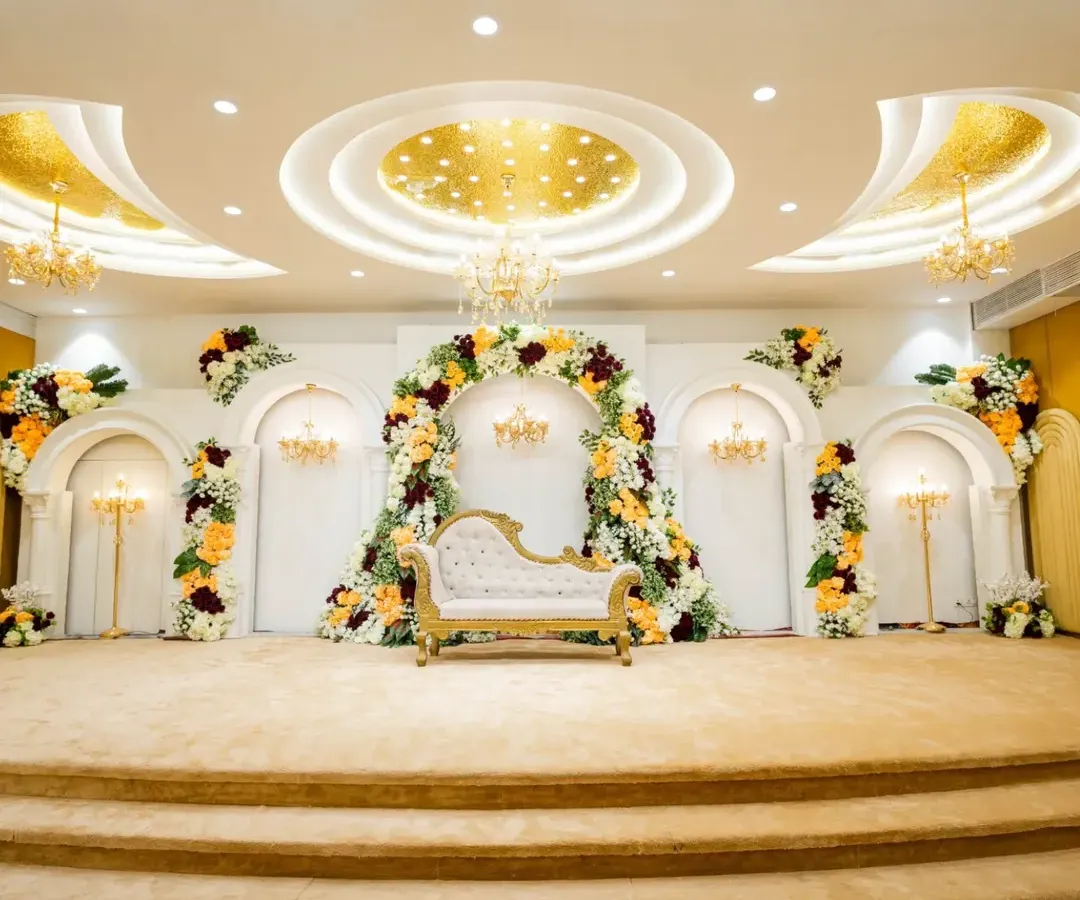This is a classic and elegant wedding stage, featuring a beautiful floral arch with rich colors and a royal diwan-style sofa. The design combines traditional aesthetics with luxurious touches like mini chandeliers, perfect for a timeless wedding ceremony.