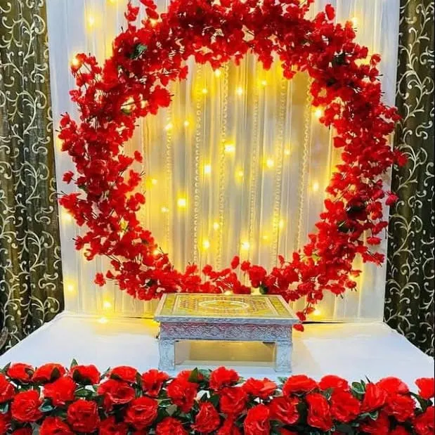 This modern Ganpati decoration features a bold circular ring completely covered in vibrant red artificial flowers. Set against a curtain of fairy lights, this design provides a contemporary and striking focal point for your celebration.