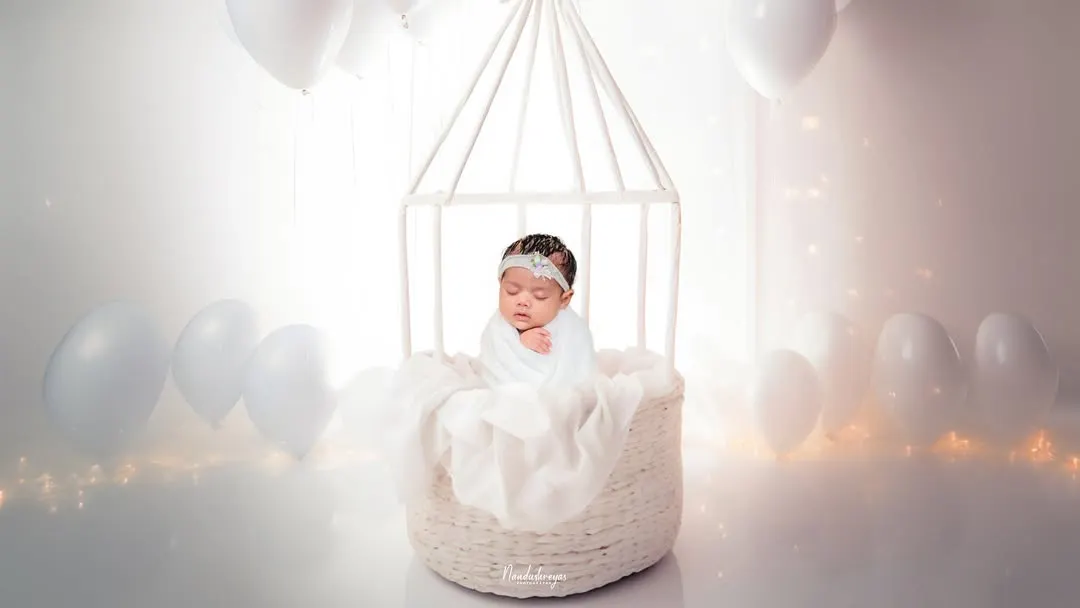 An all-white, dreamy setup to create a truly angelic portrait. Using soft textures, gentle lighting, and ethereal props like this birdcage basket, I craft a serene and timeless image of your sleeping newborn.