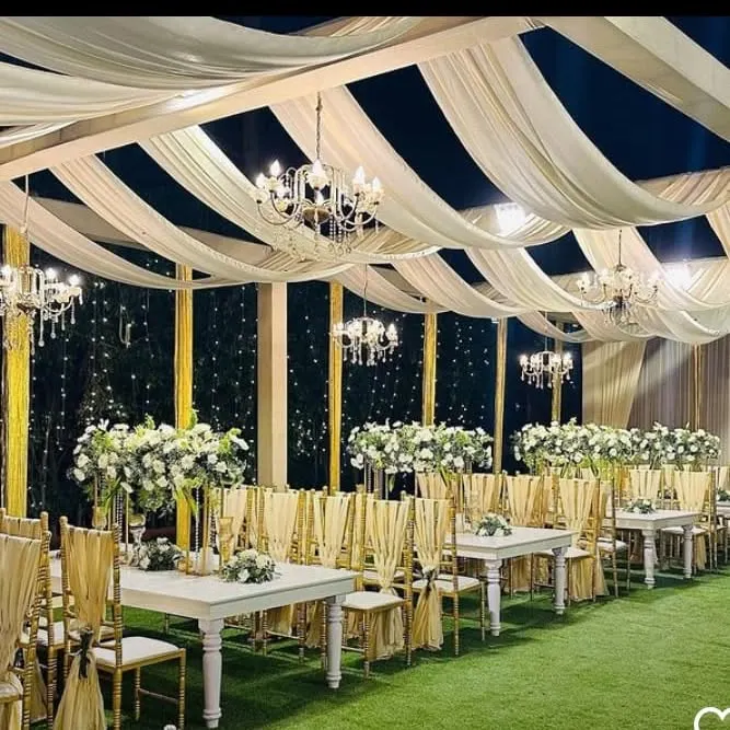 For an elegant evening reception, I arranged this outdoor setting with a fully draped ceiling, classic chandeliers, and white floral centerpieces. The setup includes white tables and gold-accented chairs on an artificial grass lawn.