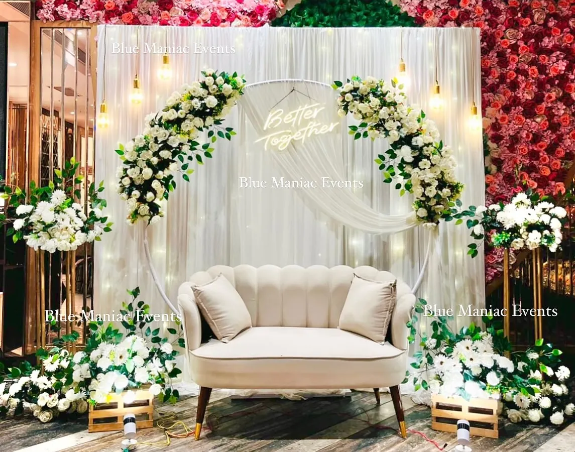 For a sophisticated and timeless celebration, we designed this beautiful white-themed Roka setup. It features a circular arch with white and pink floral arrangements, a 'Better Together' neon sign, and elegant lighting against a backdrop of red and green flower walls.