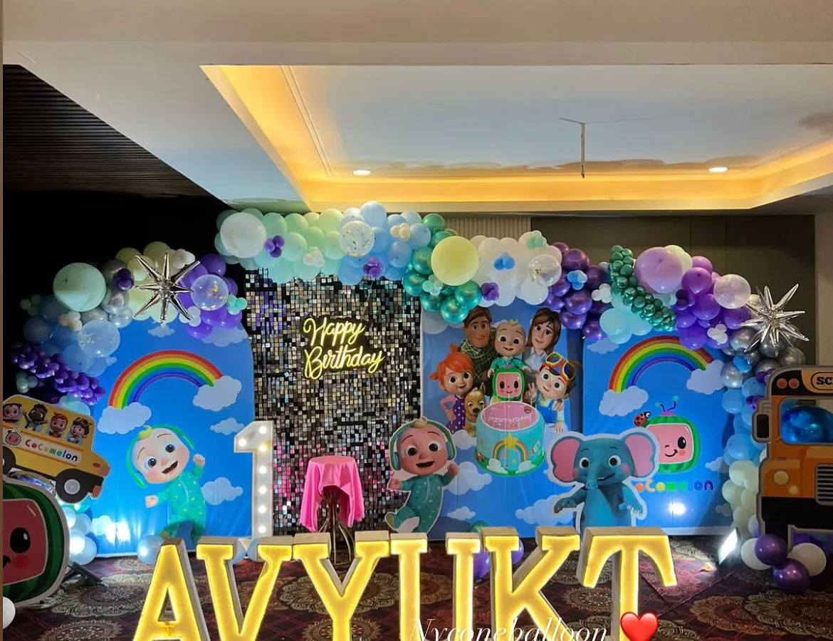 For this grand first birthday, I created a full Cocomelon wonderland. The setup includes a massive backdrop with characters, a silver sequin panel, a balloon arch, large light-up marquee letters spelling the name 'AVYUKT', and multiple character cutouts.