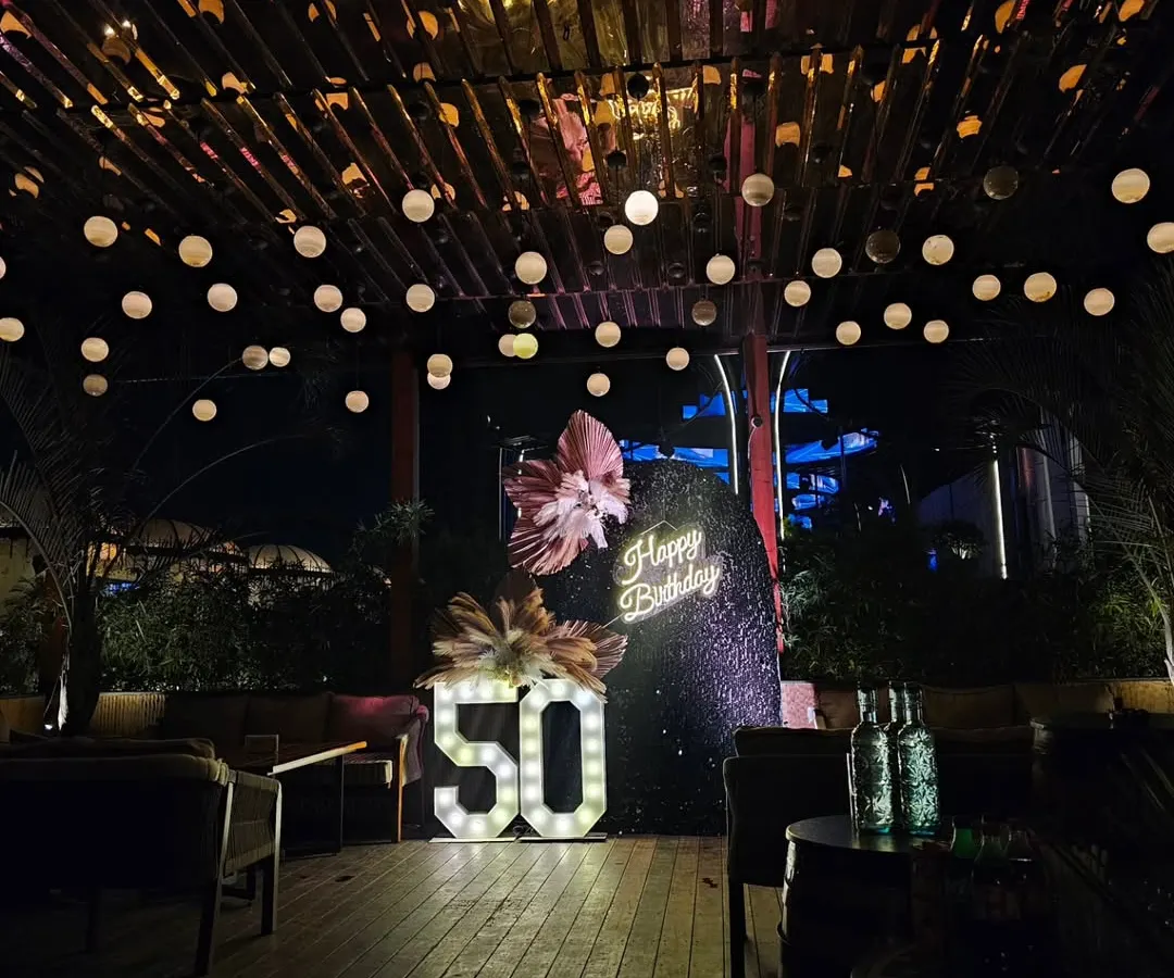 Celebrating a 50th birthday calls for something truly special. For this milestone party, I designed a sophisticated outdoor lounge setting with a dramatic sequin backdrop, a custom neon sign, and large marquee numbers, all under a canopy of warm string lights.