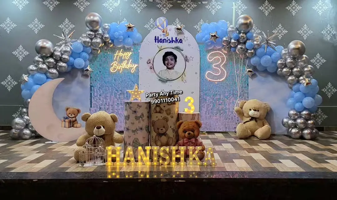 I create magical teddy bear themed parties perfect for a young child's birthday. This setup features a shimmer wall, a moon prop with a teddy, multiple teddy bears, custom cake stands, and a blue and silver balloon arch, complete with a neon birthday sign.