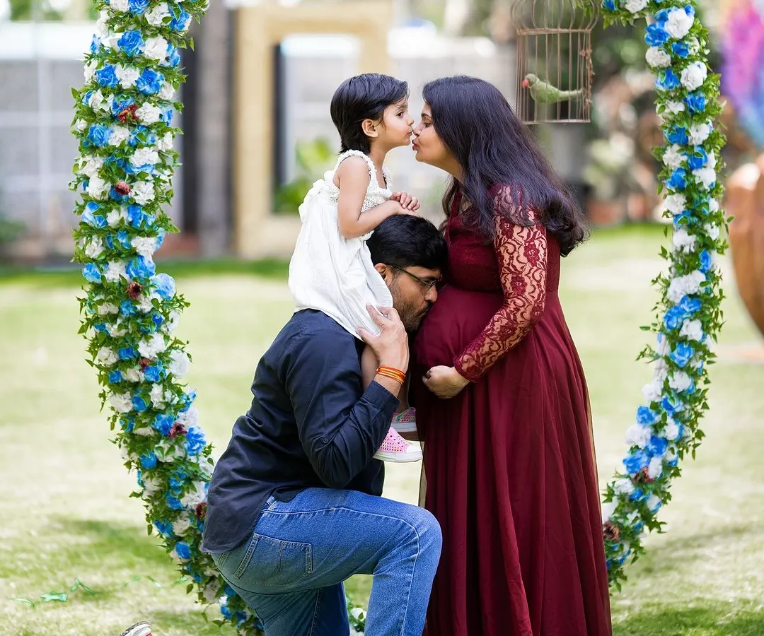 A heartwarming family maternity photo capturing a beautiful moment of connection. The older sibling shares a kiss with mom while dad listens to the baby bump, all framed by a lovely floral arch.