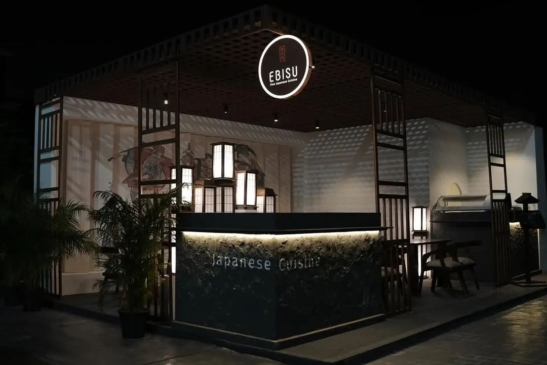 A wide shot of the 'Ebisu' Japanese fine-dining pop-up we created for the India Art Fair. The design features a structured, dark wood frame, a glowing circular brand sign, and traditional Japanese lanterns to create an authentic and inviting atmosphere at night.