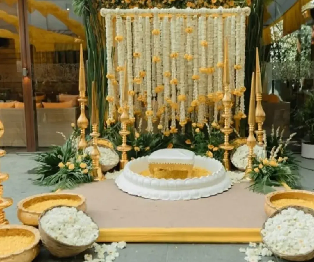 This is an elegant and traditional Haldi ceremony setup. It features a central white seat for the rituals, surrounded by bowls of fresh flower petals, all set against a beautiful backdrop of white and yellow flower strings.
