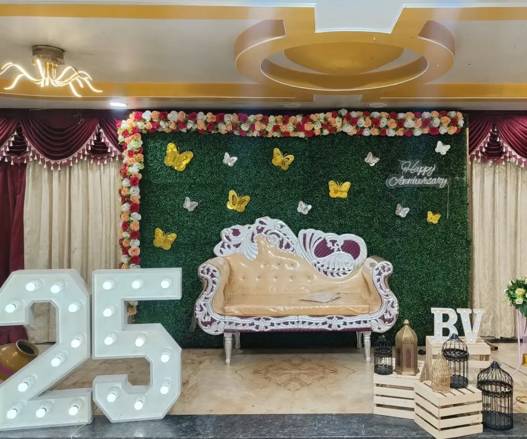 For grand milestone celebrations like a 25th anniversary, I design impressive stage setups. This features giant light-up marquee numbers, a lush green wall backdrop for photos, and an ornate sofa, turning any venue into a spectacular party space.