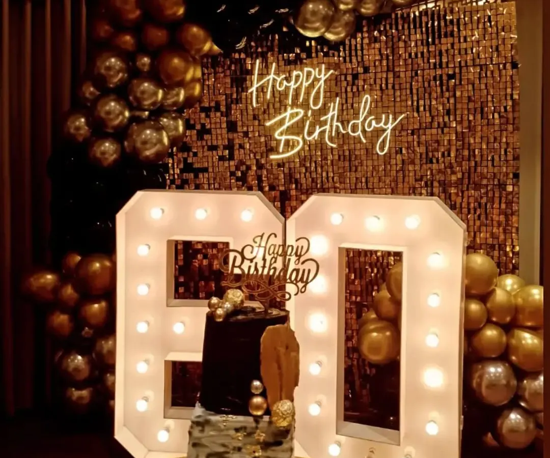 For a glamorous 60th birthday, I created this stunning photo corner. It features a gold sequin shimmer wall, a custom 'Happy Birthday' neon sign, and large marquee light-up numbers, all framed by a rich black and gold balloon garland.