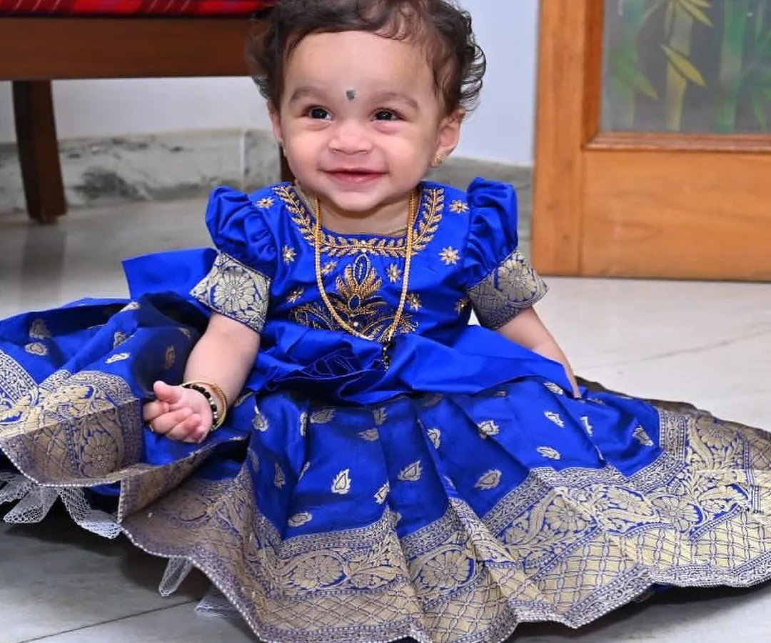 A smiling little one in her bespoke Pattu Pavadai. This royal blue silk dress, adorned with traditional gold zari work and puffed sleeves, is designed for both beauty and comfort, making it ideal for cultural ceremonies, festivals, and first birthdays.