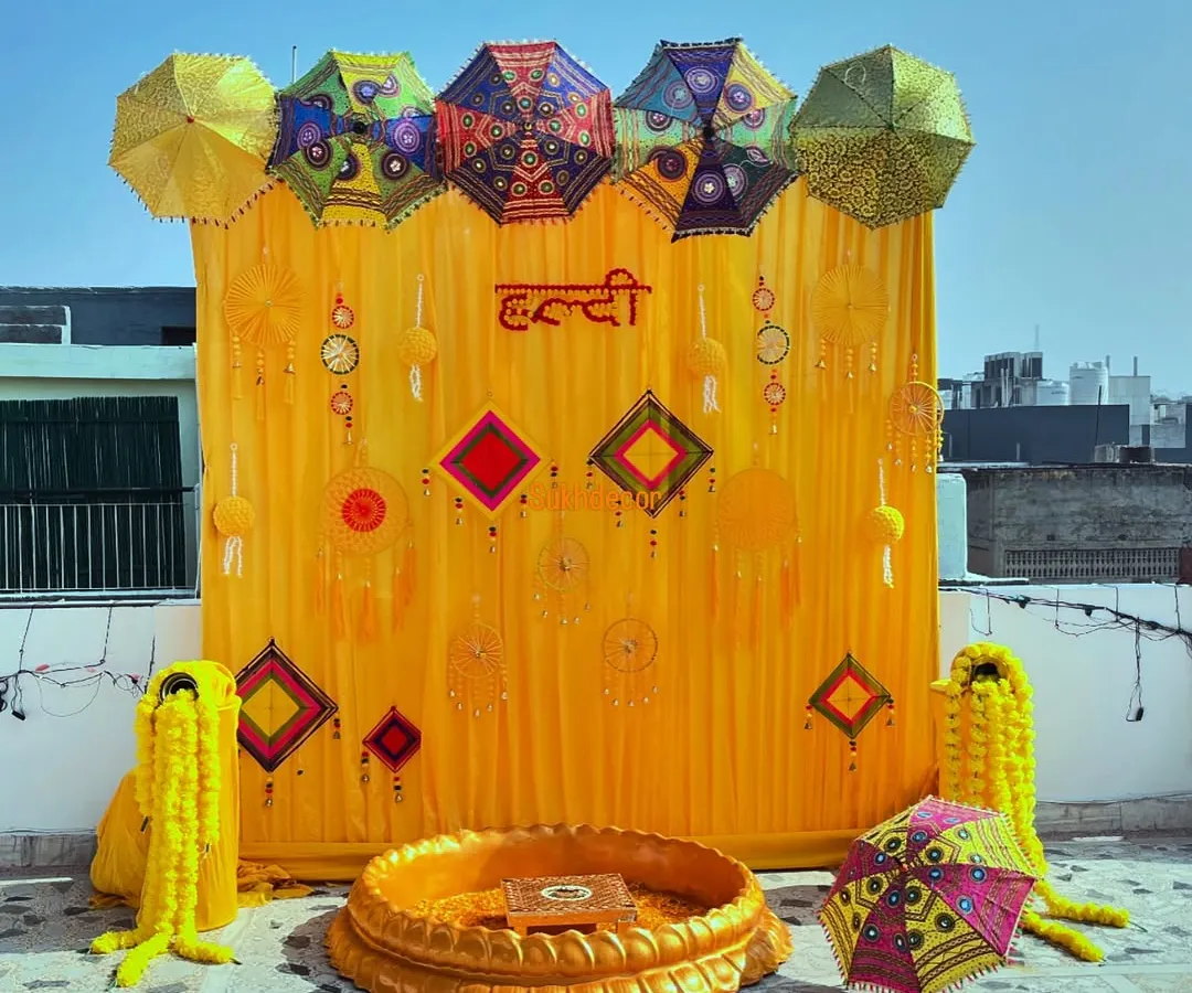 This is a classic Haldi ceremony setup I did on a sunny rooftop. The backdrop features bright yellow drapes with the word 'Haldi' in Hindi, decorated with colorful Rajasthani umbrellas and traditional hangings for a festive look.