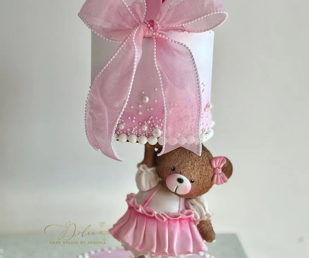 Meet one of our most loved gravity-defying designs. This adorable teddy bear, dressed in a handmade pink frock, holds up a cake for a little one's first birthday. Every detail, from the tiny bow to the delicate pearls, is crafted by hand to make your celebration extra special.