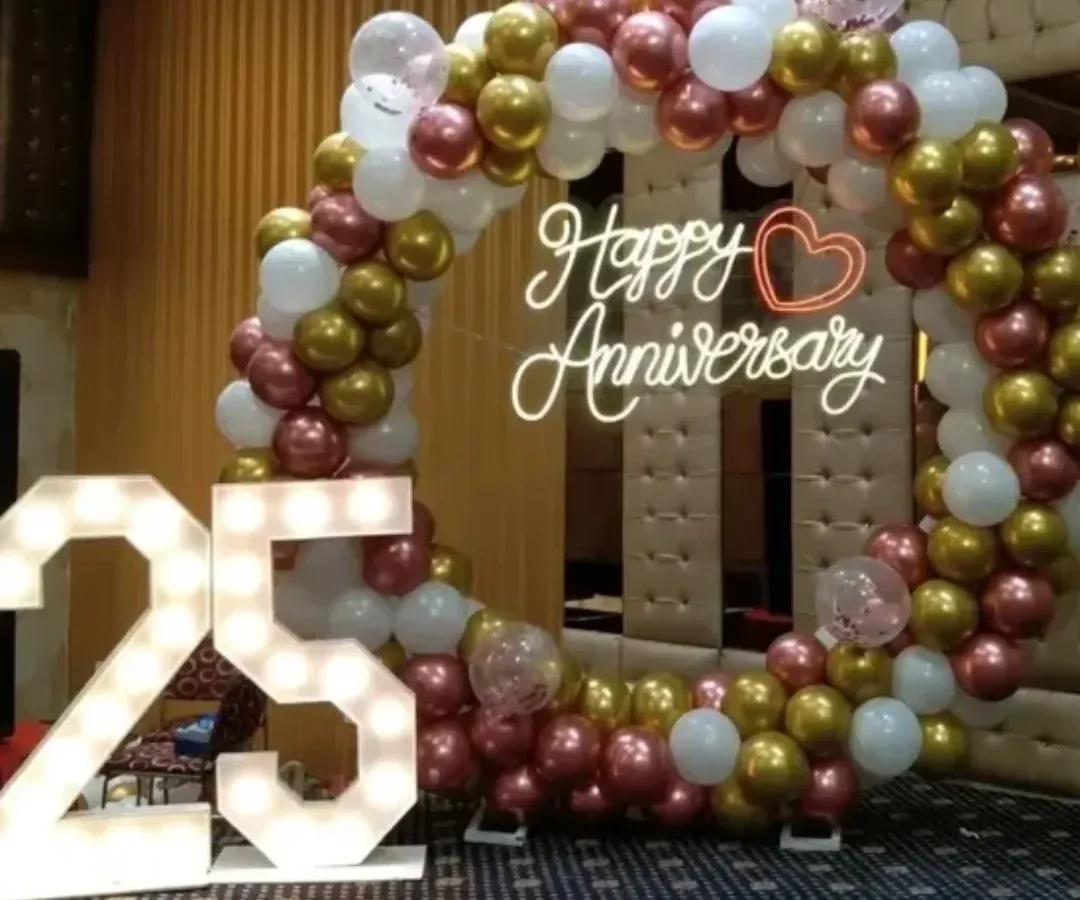 Celebrating a 25th anniversary with style. This decor includes a circular balloon arch in rose gold, gold, and white, a 'Happy Anniversary' neon sign, and large marquee numbers, perfect for creating a memorable photo opportunity in a party hall.