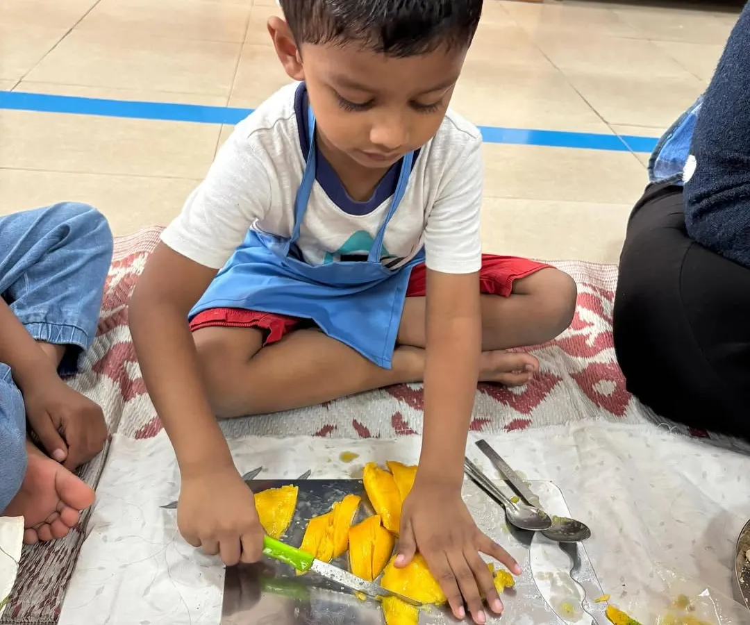 Here, a child is deeply focused on the practical life skill of cutting a mango. Using a child-safe knife, they are not just preparing a snack; they are developing fine motor skills, concentration, and a wonderful sense of independence.