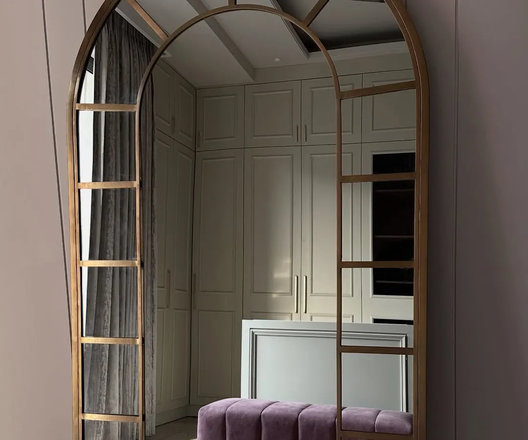 A glimpse into a walk-in closet that redefines personal luxury. This space features custom off-white cabinetry, a central island, a plush purple bench for seating, and a statement arched mirror, creating a functional and elegant dressing area.