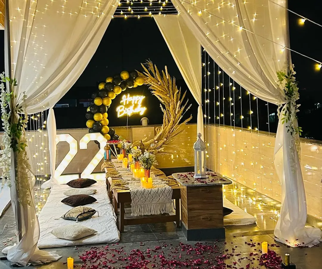 A complete view of my signature terrace cabana dinner setup for a 22nd birthday. This romantic arrangement includes a canopy with sheer white drapes and fairy lights, low seating with cushions, and rose petals scattered on the floor.