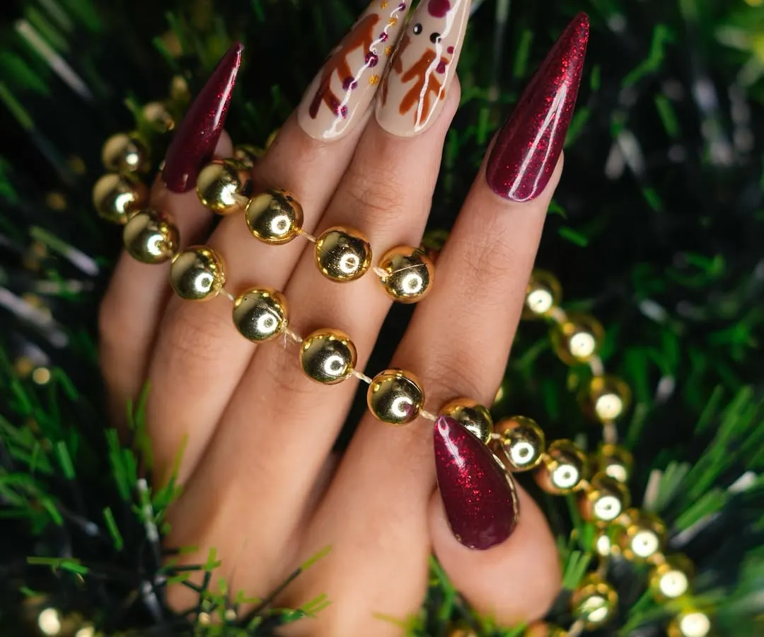 A festive Christmas look featuring sculpted stiletto extensions. This set combines deep red glitter nails with nude accent nails, which are detailed with hand-painted reindeer art for a touch of holiday magic.