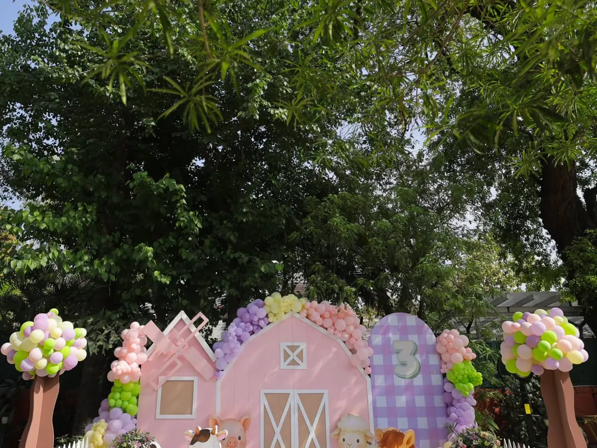For this third birthday, we designed a dreamy pastel barnyard in an open garden. This wide view showcases the bespoke pink barnhouse backdrop, complete with adorable animal props and whimsical balloon trees, creating a picturesque countryside celebration.