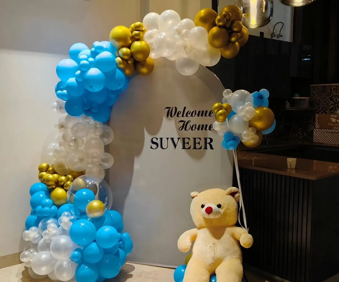 For a modern and clean 'Welcome Home' celebration, I created this setup for baby Suveer. It features a stylish blue, white, and gold balloon arch against a simple white circular backdrop, complemented by a cute teddy bear.