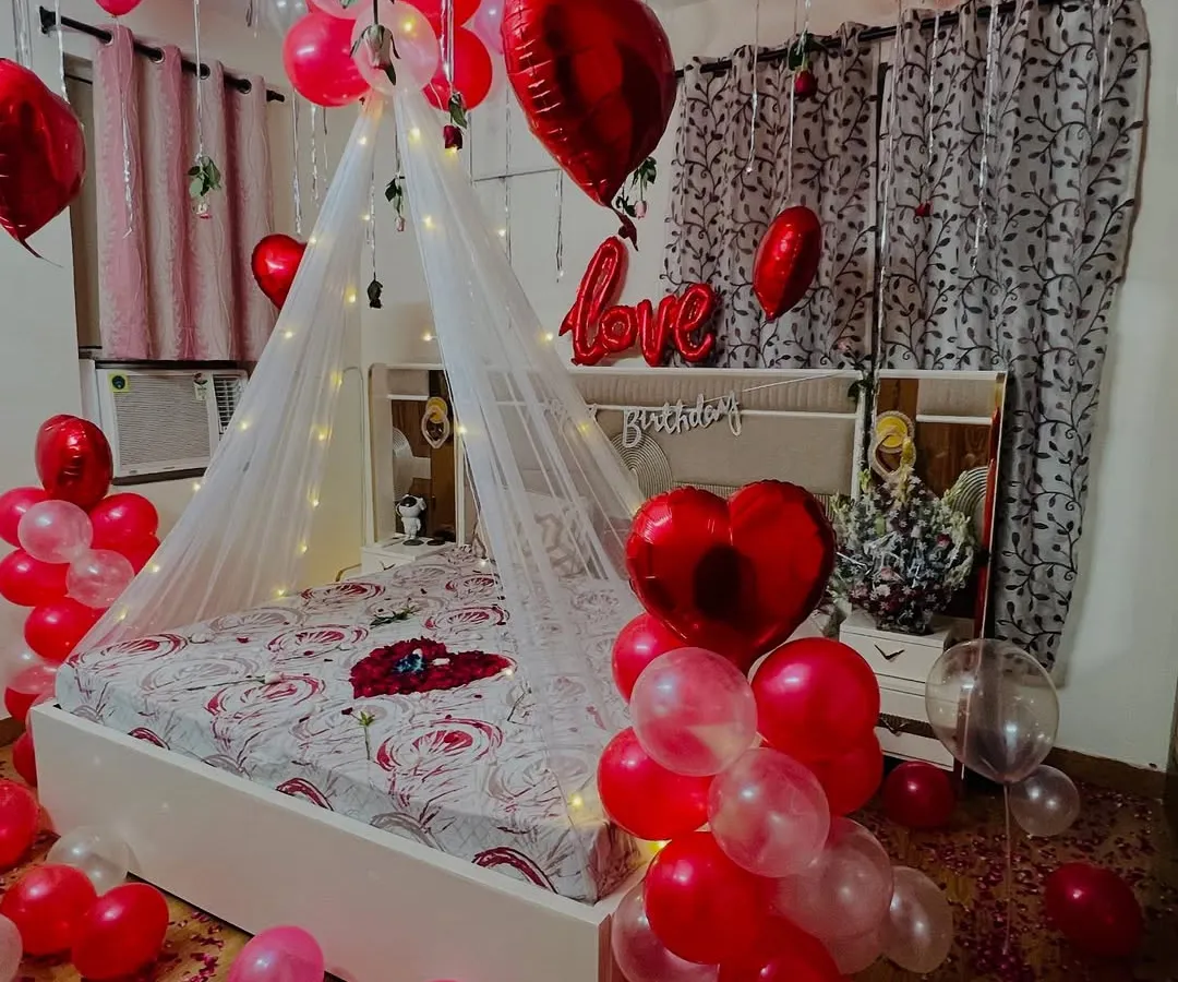 I design romantic room surprises for anniversaries and special moments. This setup features a fairy light canopy over the bed, with red and white balloons, heart-shaped foil balloons, and rose petals to create a truly memorable experience.