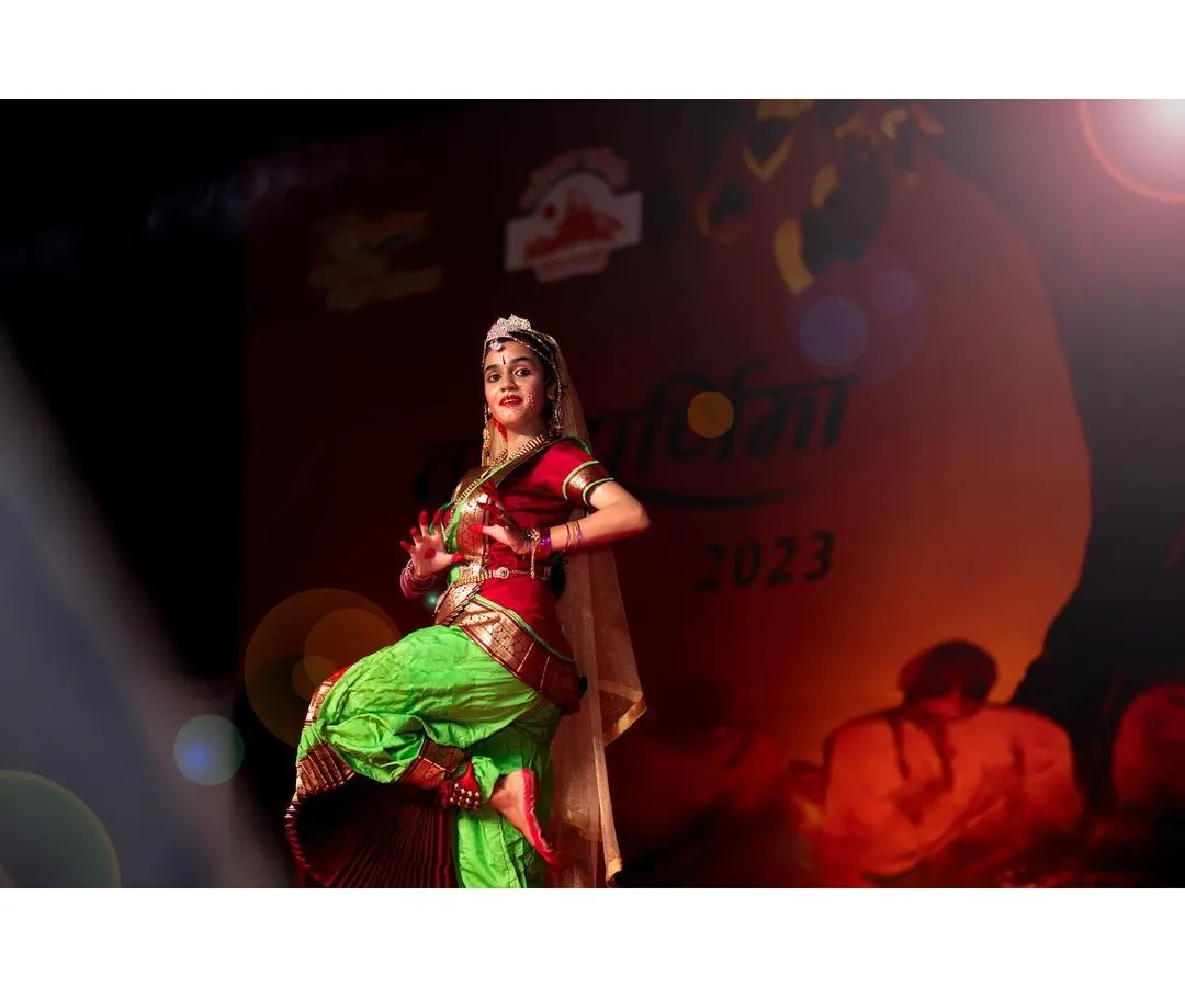 A Bharatanatyam dancer strikes a powerful pose on stage, her vibrant costume caught in the dramatic stage lighting. I focus on freezing these peak moments of expression and movement during live classical dance performances.