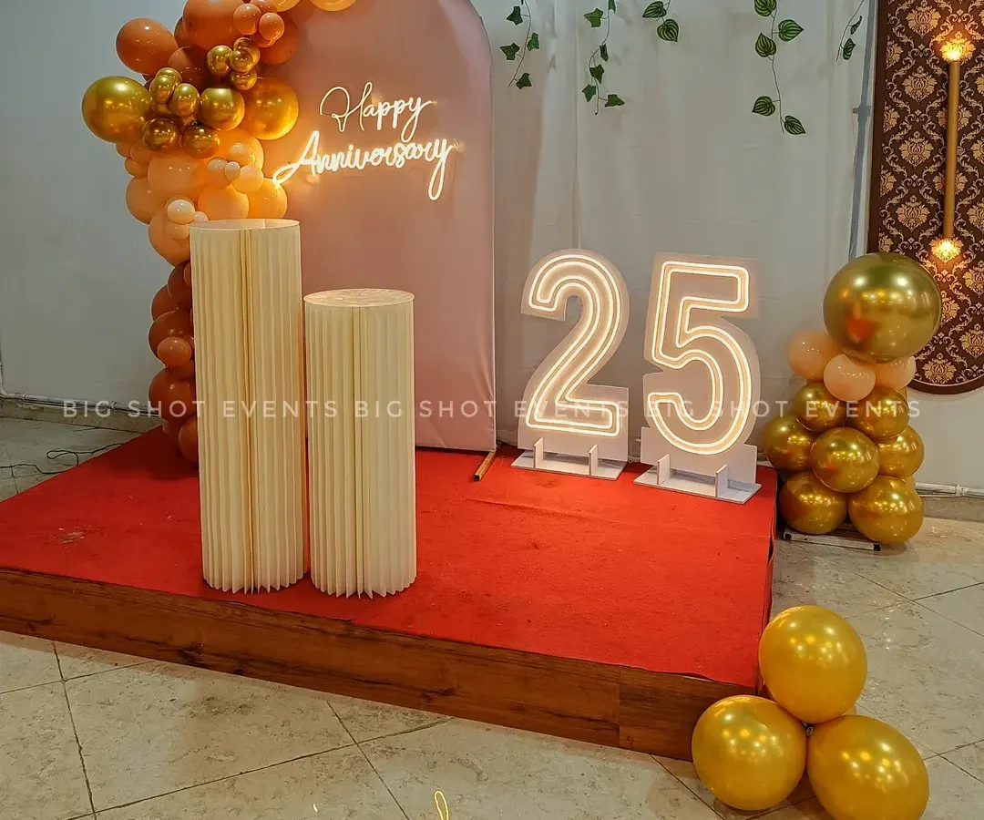 For this 25th anniversary, we designed an elegant stage with a 'Happy Anniversary' neon sign, large light-up marquee numbers, and a stylish balloon arch in a sophisticated gold and peach color scheme.