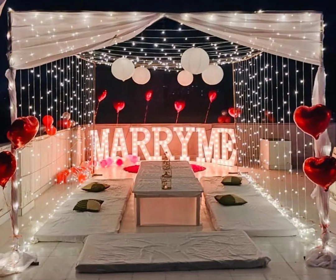 I designed this romantic rooftop proposal within a fairy light cabana. The setup includes low floor seating for an intimate dinner, heart-shaped foil balloons, and glowing 'MARRY ME' marquee letters as the centerpiece.