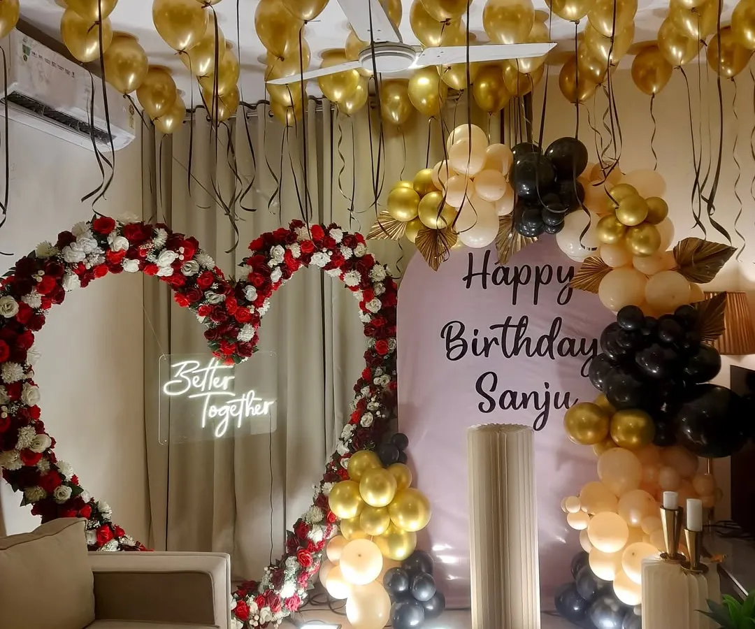 For an elegant birthday or anniversary, I combined a heart-shaped frame made of red and white roses with a stylish black, gold, and peach organic balloon garland. The "Better Together" neon sign and golden ceiling balloons complete this romantic surprise party decoration.