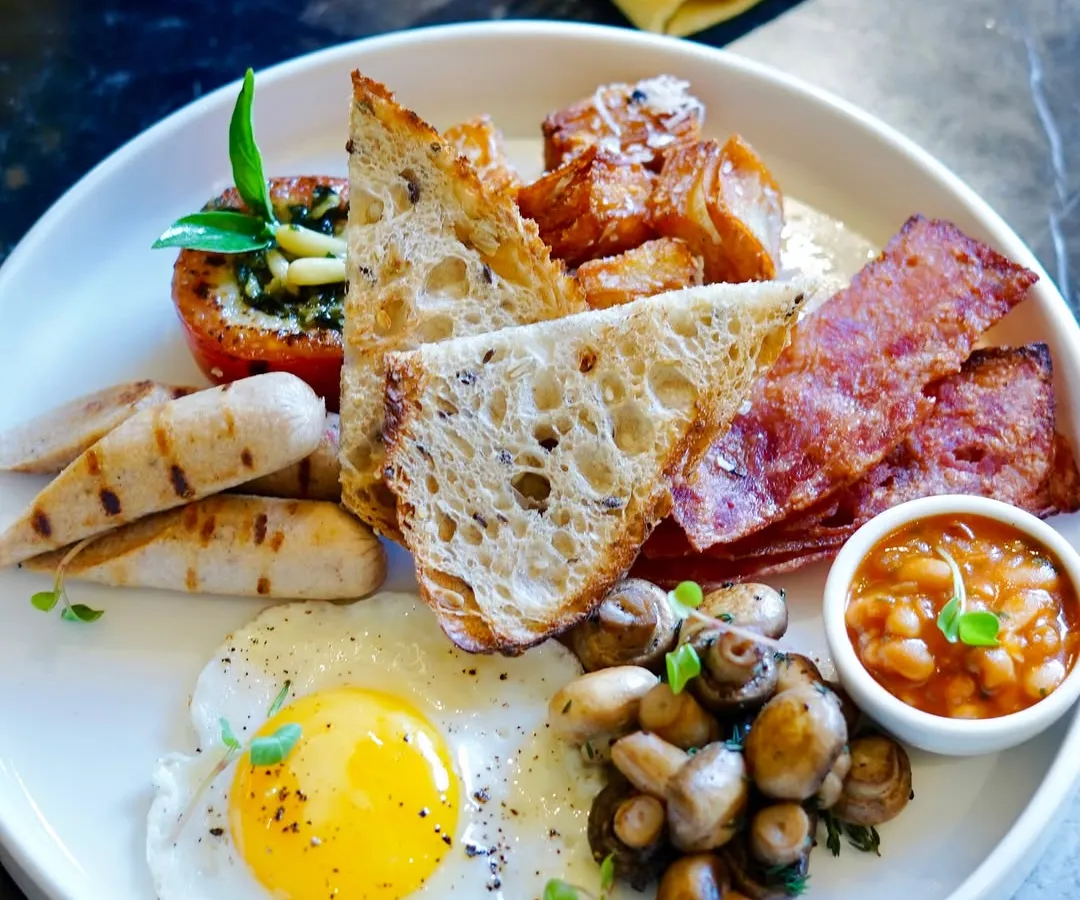 A classic full breakfast, perfectly plated and ready to enjoy. This hearty meal includes a sunny-side-up organic egg, sausages, bacon, baked beans, sautéed mushrooms, and sourdough toast, showcasing our commitment to quality ingredients for your morning meal.