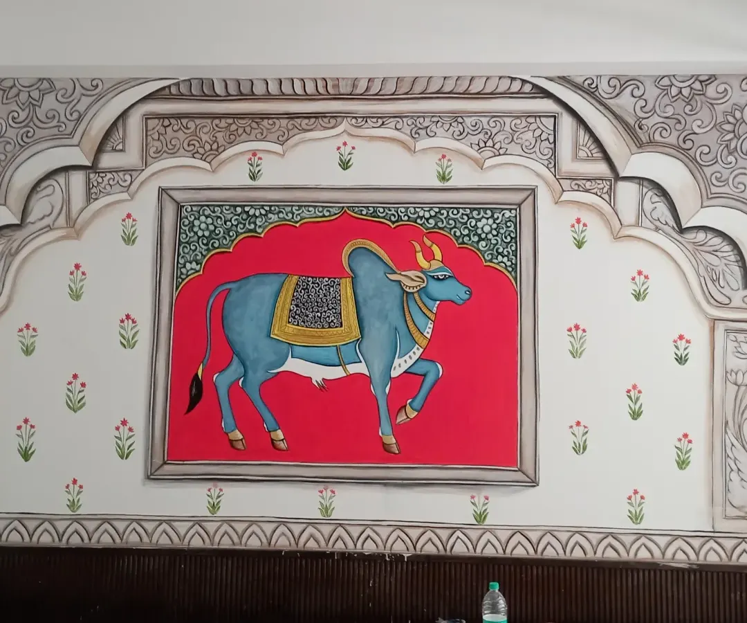 A classic Pichwai style painting, this mural brings serenity and tradition into the home. Featuring a beautifully adorned cow against a vibrant red backdrop, framed by intricate Rajasthani architectural motifs, it is a piece of heritage art that creates a divine atmosphere.