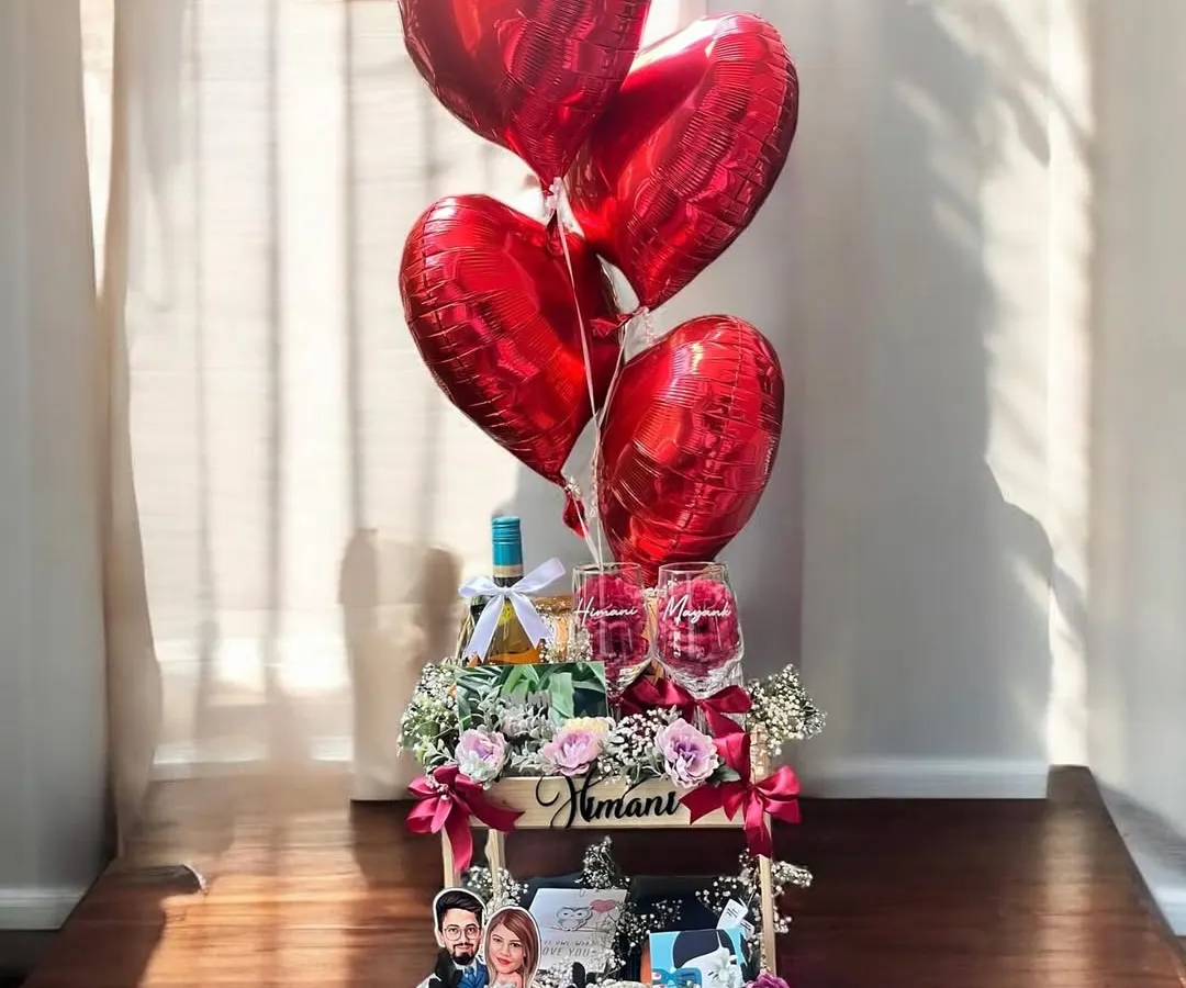 I designed this romantic anniversary hamper to be a showstopper, featuring a large wooden crate filled with flowers, personalized photo frames, wine, and elegant glasses, all topped with heart-shaped red foil balloons.