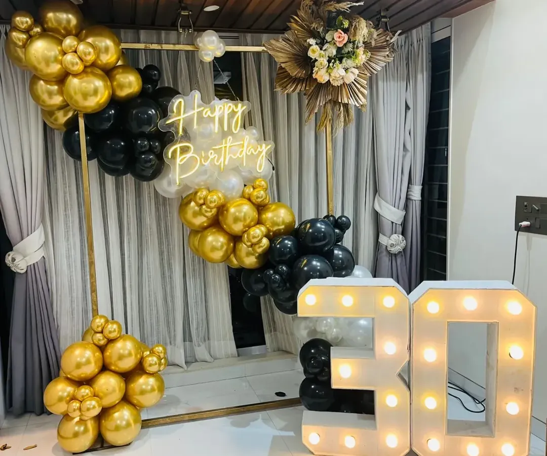 Celebrating a 30th birthday with style. This chic setup features a modern gold, black, and white balloon arrangement on a metal frame, a custom 'Happy Birthday' neon sign, and large marquee numbers, creating a sophisticated photo corner for the party.