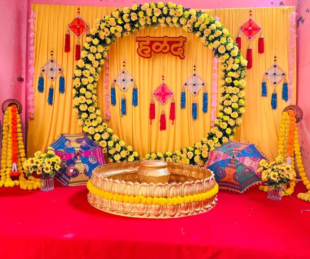 A vibrant and traditional Haldi ceremony backdrop. This setup features a stunning full circle arch made entirely of fresh yellow marigolds (genda phool) against a bright yellow curtain, with a custom 'हळद' sign in Hindi at the center.