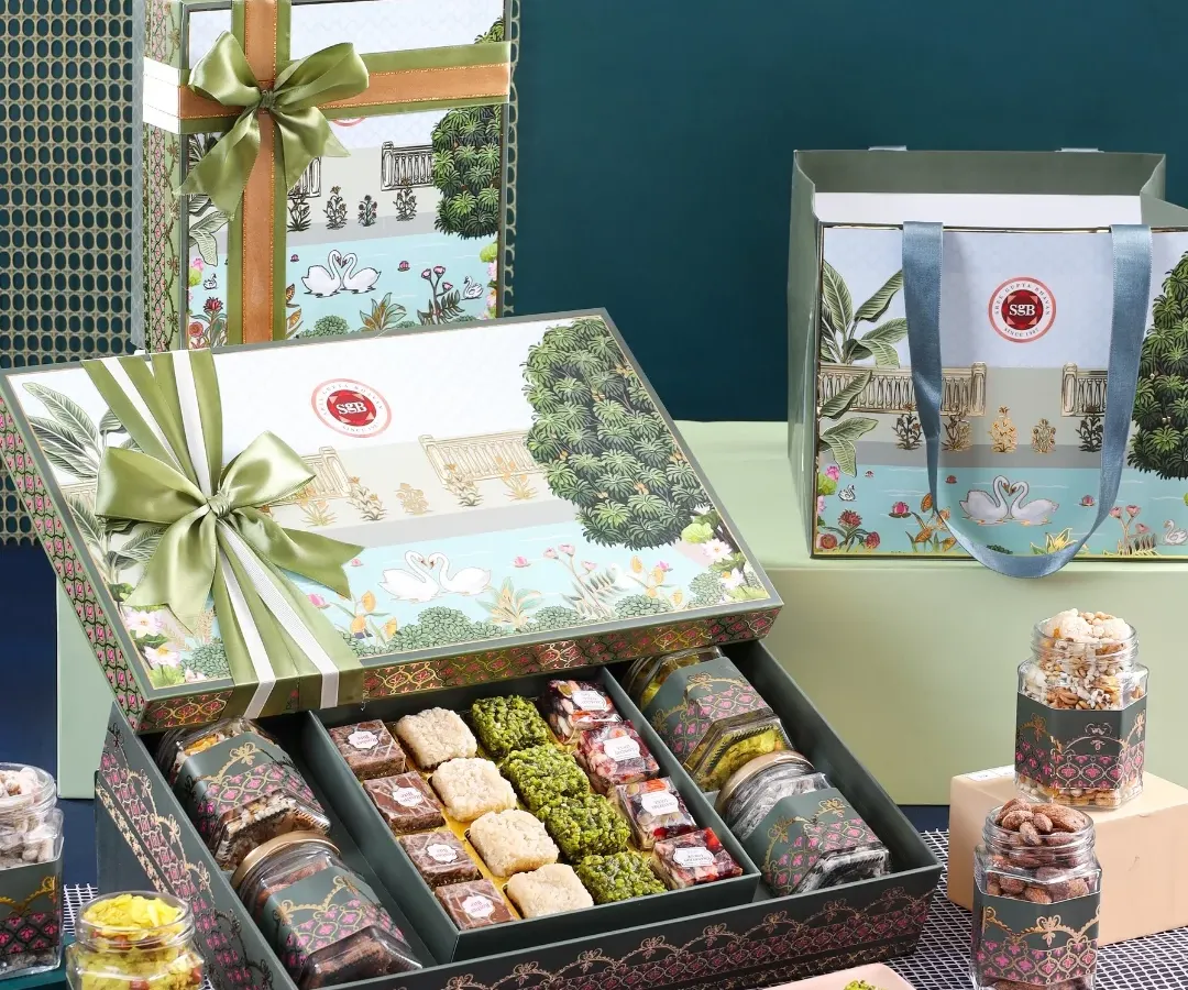 This exquisite hamper, featuring intricate illustrations and a beautiful satin bow, blends wellness and indulgence. Inside, a curated selection of gourmet sweets and premium dry fruits awaits, making it a perfect gift for any festive occasion.