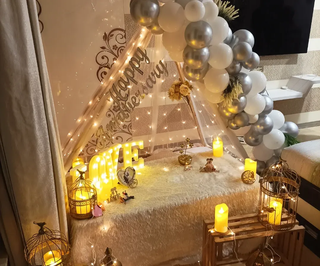 For a truly romantic anniversary, I can create an intimate cabana tent right in your home. This setup includes a plush rug, soft lighting from fairy lights and lanterns, and a beautiful silver and white balloon arch.
