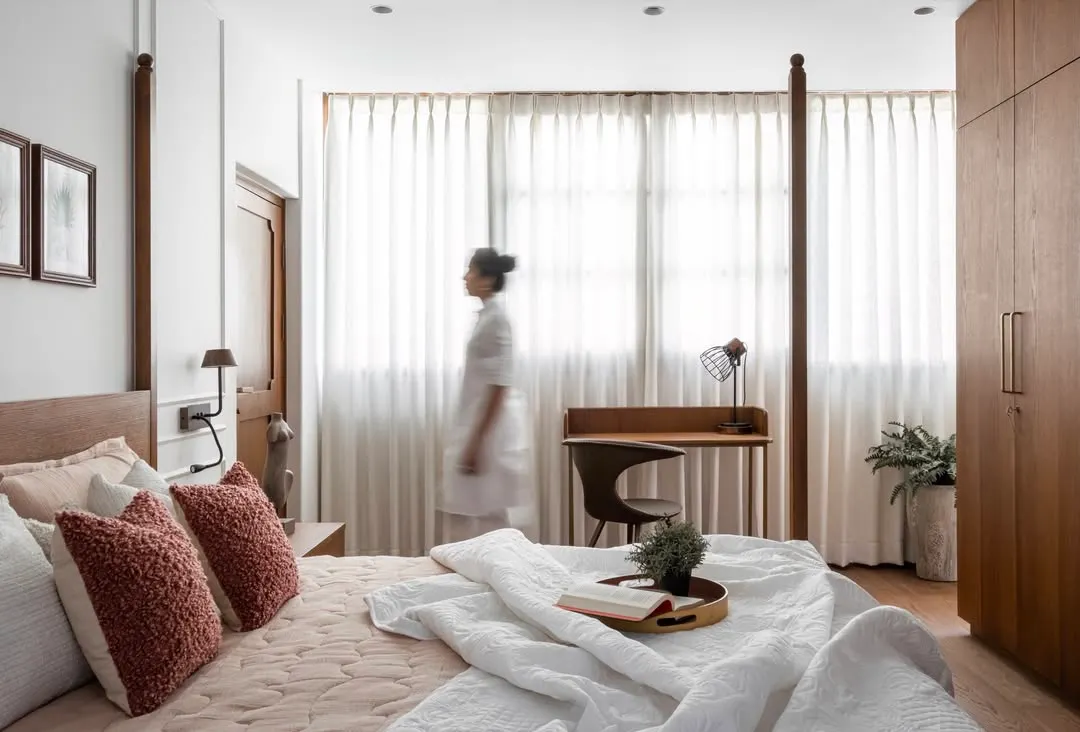 In the master bedroom, a grand four-poster bed nods to Kerala’s architectural roots, bringing its influence full circle. The rest of the space unfolds in quiet gestures: sunlight diffused through sheer drapes, soft rust against layers of white, and warm wood meeting pale walls. Nothing shouts, nothing jars; everything here is in service of stillness.