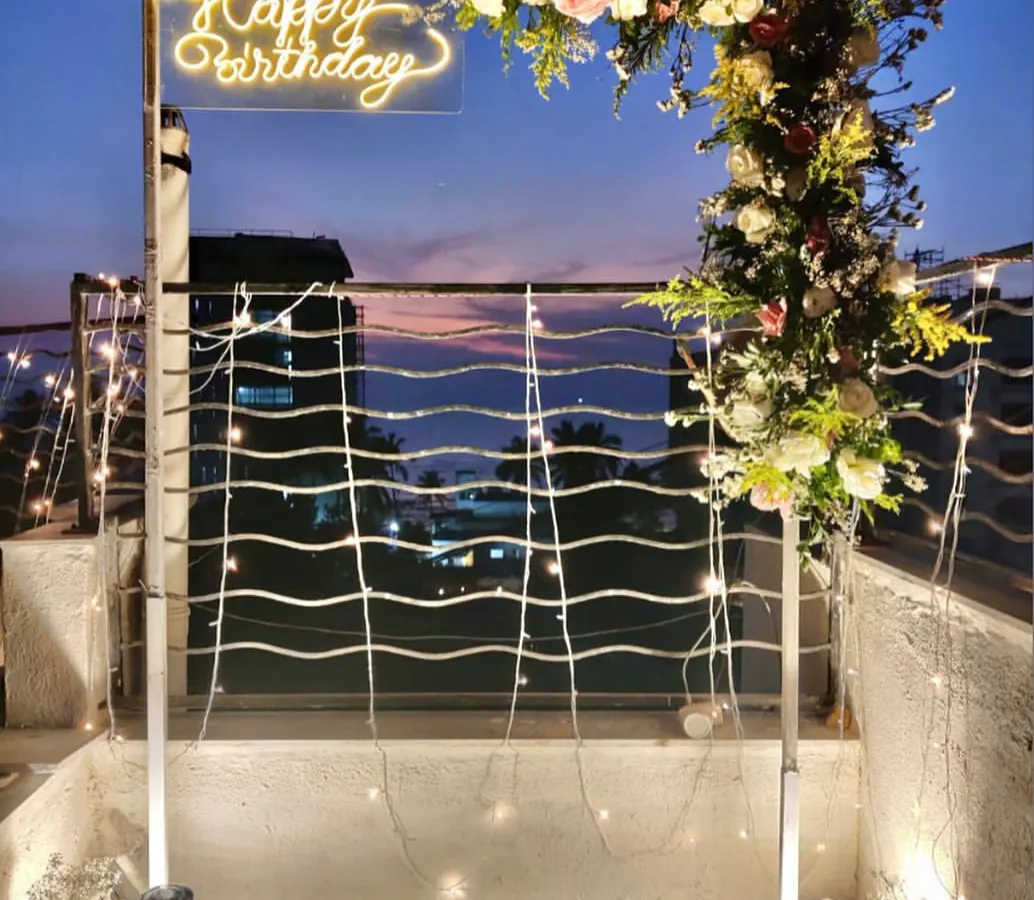 For this terrace birthday party, I created a modern and chic atmosphere with a custom neon "Happy Birthday" sign on a floral arch, set against a beautiful twilight city view. The setup includes fairy lights and lanterns for a warm, inviting glow.
