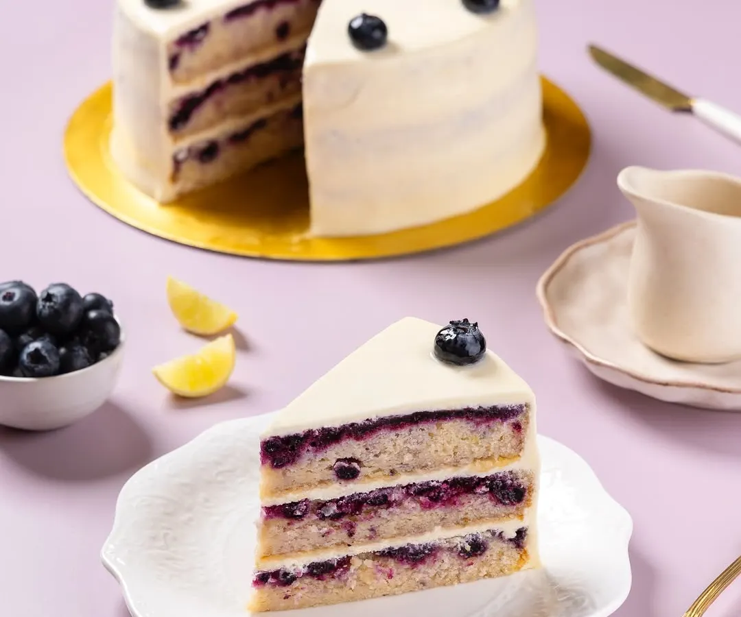 A slice of a multi-layered blueberry and lemon cake, photographed for The Big Chill Cafe. The focus is on revealing the cake's internal structure, showing the fresh fruit filling and soft sponge, a key shot for any bakery or dessert menu.
