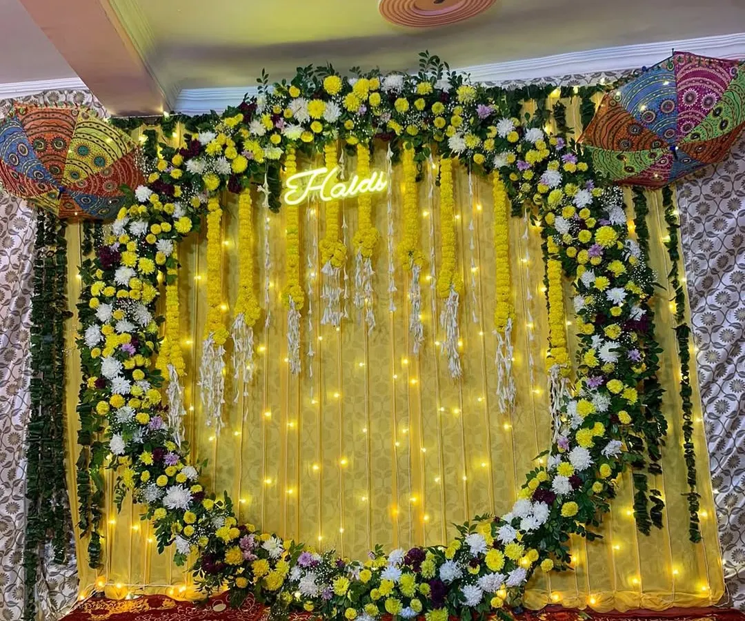A vibrant and modern Haldi backdrop. This setup features a full circular arch covered in yellow and white flowers, with a custom 'Haldi' neon sign glowing in the center against a bright yellow drape and fairy lights.