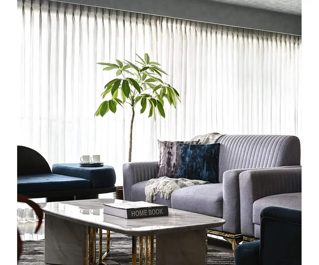 A serene living room where a neutral grey palette is enlivened by deep blue accents. The space is unified by luxurious materials, including a marble coffee table with brass legs and plush, fluted sofas, all set against sheer curtains that bathe the room in soft, natural light.