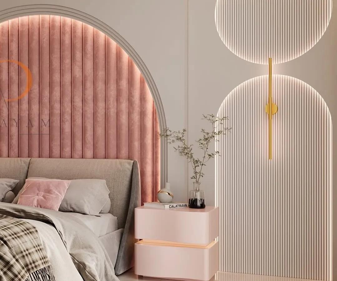 A modern bedroom design featuring a striking arched headboard with pink velvet channel tufting. The space is enhanced by custom backlit wall panels and a matching bedside table, creating a soft, luxurious, and contemporary ambiance.