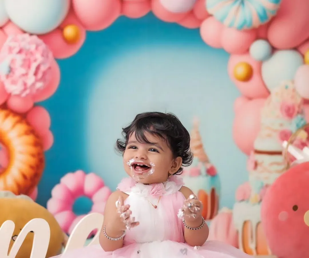 The pure joy of a first birthday cakesmash. This little girl's happy, messy smile is captured against a vibrant candy-themed backdrop with pink and blue balloons, showing a fully styled milestone session.