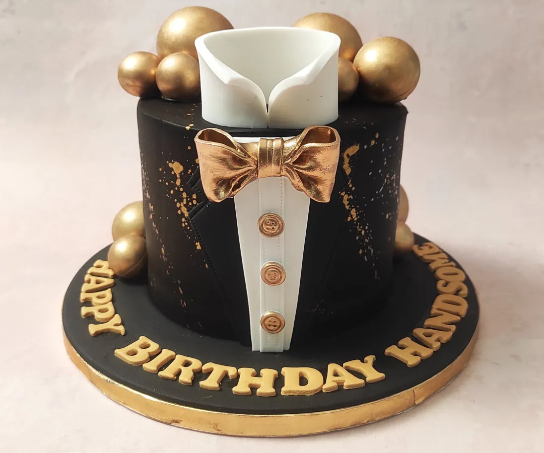 We present the essence of formal sophistication with our black and gold tuxedo cake. This design features a sharp, edible shirt collar and a lustrous gold bow tie set against a matte black tier, accented with gold splatters and spheres, making it an impeccable choice for a gentleman's milestone birthday.